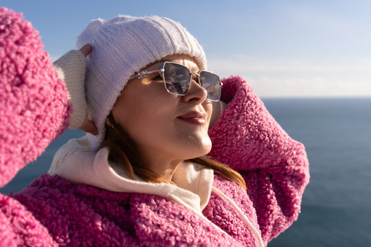 Young woman in pink fleece jacket and beanie enjoys sunny weather standing near calm ocean during winter day