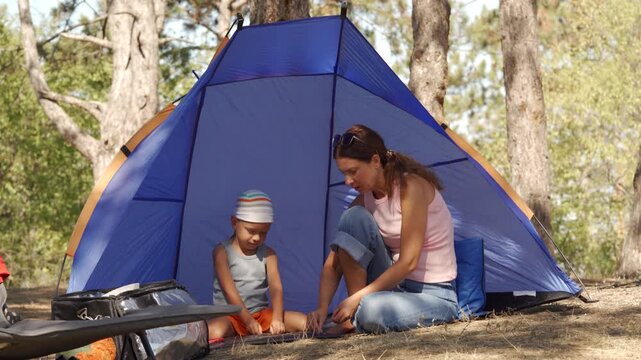 A happy mother and son sit together inside their blue tent, playing quietly. They feel content and bonded while relaxing in the sunny pine forest.