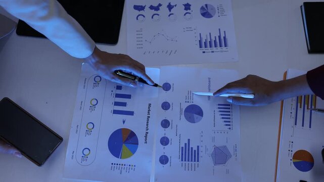 Top view of business hands pointing at financial charts, graphs, data analytics, and performance metrics during a corporate meeting or strategy presentation.
