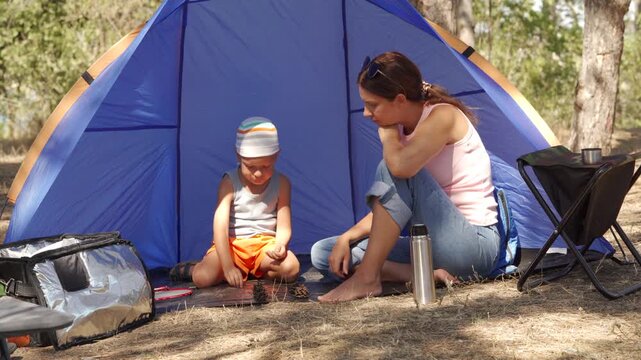 A mother and son sit together inside a blue tent at a forest campsite. They play quietly with pinecones during a peaceful trip. The happy moment feels calm and full of love.