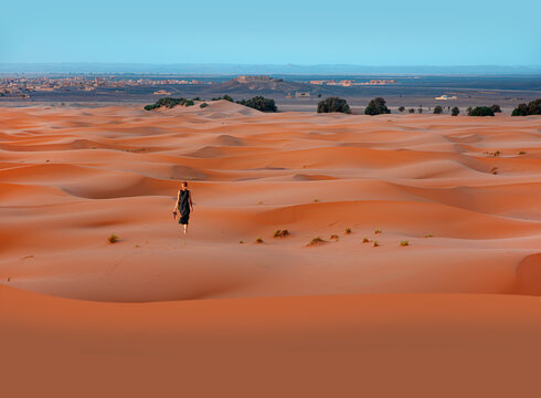 A young blonde woman in a long black dress walking alone in the Sahara Desert - General view of the Merzouga hotels district - Morocco, Africa