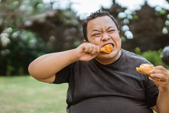 portrait of overweight man eating fried chicken hungrily from two hands against an outdoor background