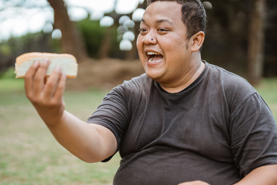 overweight young man mouths while eating bread held in his hand with an outdoor background