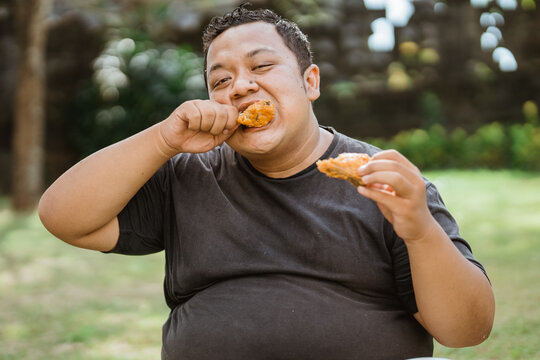 portrait of overweight Asian man eating fried chicken from two hands against an outdoor background