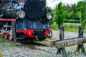 Vintage black steam locomotive with red details and large headlights, standing on railway tracks behind a buffer stop, close-up view of the engine front, green trees background, outdoor daylight. © Adam