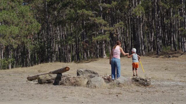 A mother and her young son hold hands while standing on rocks near a pine forest. She gently guides him during their sunny walk. The peaceful moment feels safe and full of love.