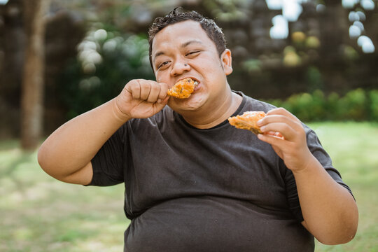 fat asian man eating fried chicken from two hands against an outdoor background