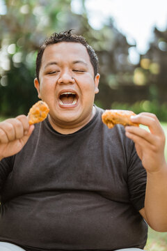 close up of a fat man holding a chicken drumstick and opening his mouth wide while eating with outdoor background