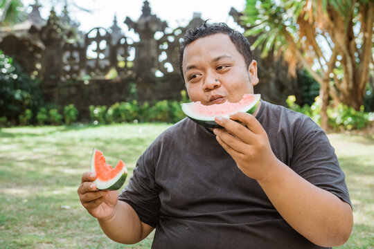 fat man eating and holding watermelon slices with two hands in outdoor background