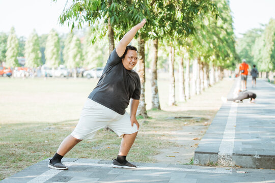 fat man smiling while doing side bends stretching arm and thigh muscles before exercising in park