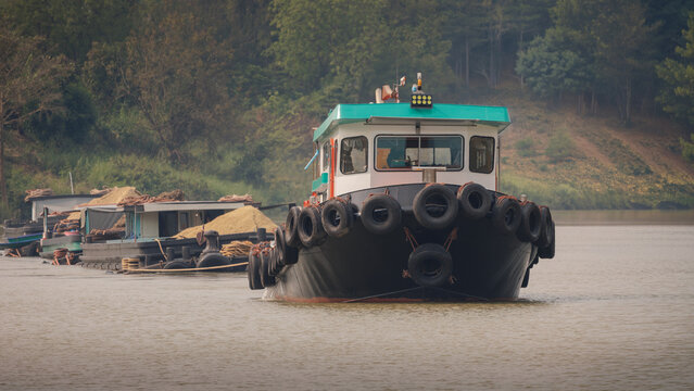 tugboat navigating along river, shot head-on, forested shoreline in background, vessel equipped with numerous used tires as protective fenders around hull, river transportation, commercial waterway