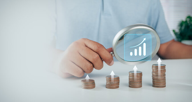 Person holding magnifying glass inspecting growing coin stacks with digital bar chart and upward arrow hologram icon, concept of financial investment growth, profit analysis, and economic success.
