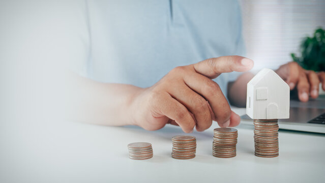 Person pointing at growing coin stacks steps with wooden house model on top, concept of saving money for house purchase, property investment growth, mortgage planning, and financial success.