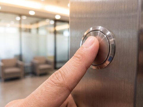 A close-up shot of a person's finger pressing a shiny metallic elevator call button in a modern office building.