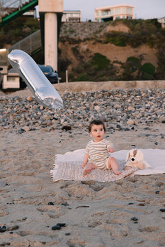 Curious baby sits on a beach blanket with a birthday balloon