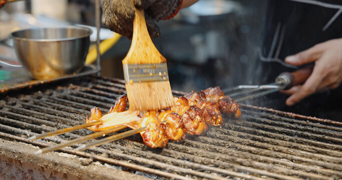 Chef brushing sauce on meat skewers, grilling on outdoor street food stall, Asian night market culinary experience, professional cooking process.