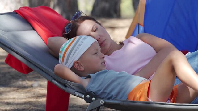 A loving mother and her small son lie on a folding lounger together at a campsite. They rest peacefully under shady trees. They feel calm, cozy, and happy.