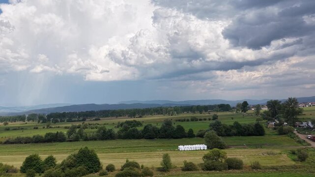 Rural fields and tree lines extend toward distant hills beneath dramatic storm clouds with a visible rain shower on the horizon.