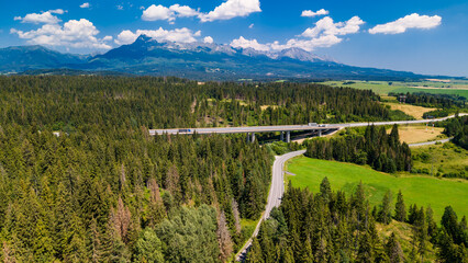 Fototapeta premium Winding road and mountains in the background during daytime. A winding road weaves through green fields and forests, mountains looming in the distance.