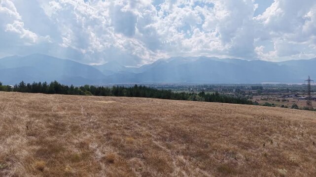 Golden grassland stretches toward a distant mountain range while the aerial camera moves slowly across the countryside beneath a dramatic cloudy sky.