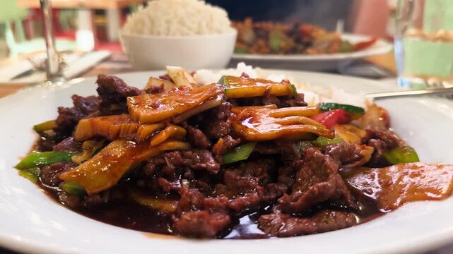 Asian-style beef stir fry with tender sliced beef, colorful vegetables and glossy savory sauce served on a white plate with steamed rice in the background.