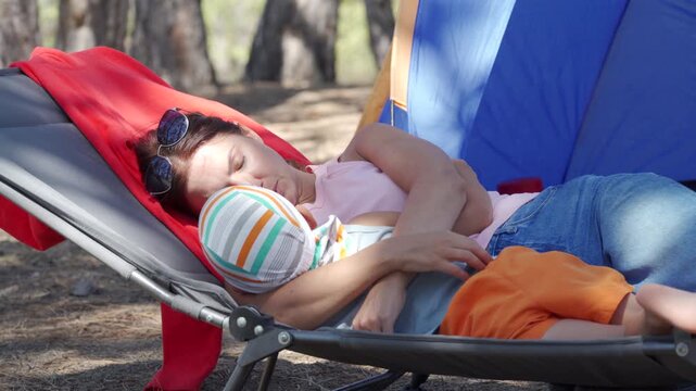 A loving mother cuddles her young son as they sleep peacefully on a folding cot. They share a tender, cozy moment beside a blue tent at a forest campsite.