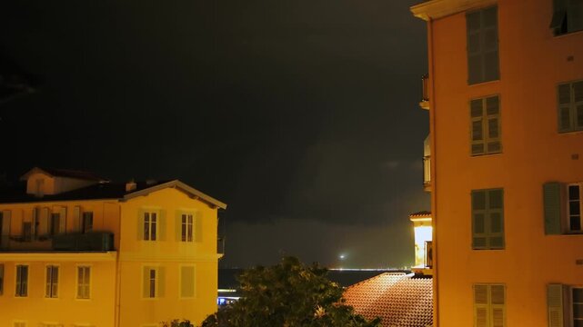 Dramatic nighttime view of a powerful thunderstorm with lightning flashing in the dark sky above a coastal city, illuminating the buildings and the distant shoreline in a spectacular display