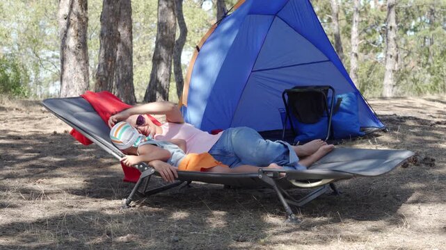 A loving mother and her small son cuddle comfortably on a camping cot. She looks at him with affection while he rests peacefully. They enjoy a calm, cozy afternoon near their tent in the forest.