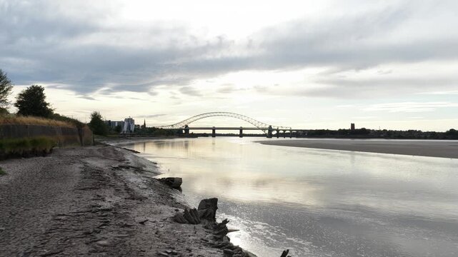 Rising tide covering muddy riverbank at sunset with bridge view