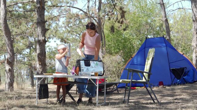 A mother and her young son unpack food containers together at a folding table. They enjoy a happy, peaceful picnic beside a blue tent in a sunny forest setting.