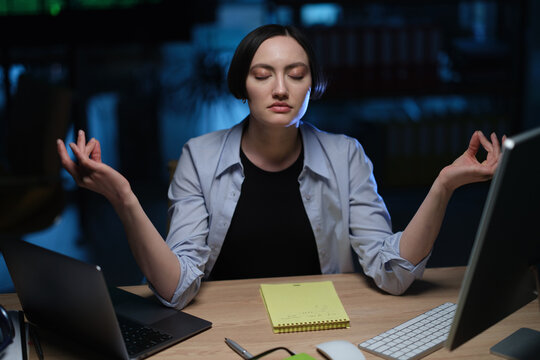 Female employee meditates with closed eyes while sitting at office desk. Woman raises hands calmly practicing relaxation during stressful work
