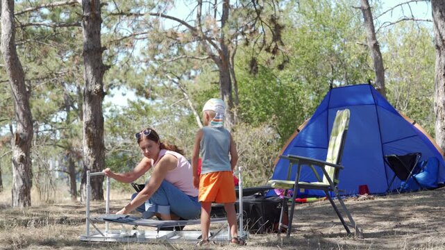 A mother sets up a folding table while her young son watches helper-style. They enjoy a peaceful, joyful camping moment near a blue tent in the sunny woods.