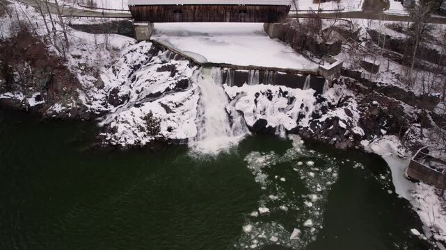 Wide aerial drone shot slowly pulling away from a historic Willard covered bridge and waterfall along the snowy Ottauquechee River near White River Junction, Vermont.