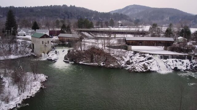 Aerial drone approach toward the Willard covered bridges, old mill and island with waterfall along the snowy Ottauquechee River, with I-91 visible in the background near White River Junction, Vermont