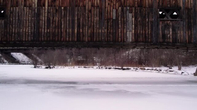 Aerial drone slowly pulling away from a historic Willard covered bridge and waterfall along the snowy, partially frozen Ottauquechee River near White River Junction, Vermont.