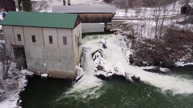 Aerial drone orbit around a Willard covered bridge, waterfall and historic mill along the snowy Ottauquechee River near White River Junction, Vermont, with partial river ice.