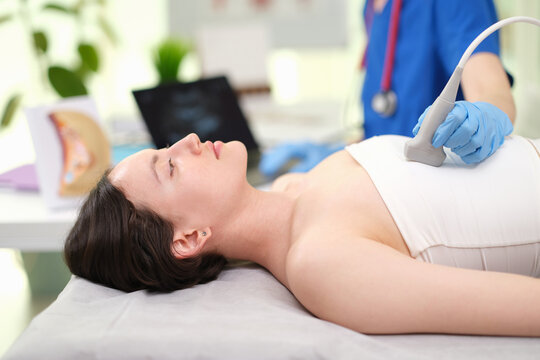 A woman is calmly undergoing an ultrasound examination of breast in a medical office setting, viewed from the side. The scene features a medical examination table, a pillow, and an ultrasound machine.