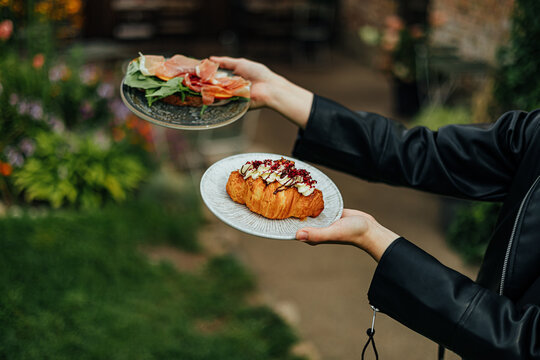 Person holds two plates with an open prosciutto and leafy green sandwich plus a cream-topped croissant, photographed outdoors with soft garden bokeh and copy space
