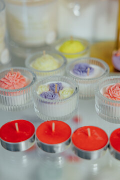 Decorative candles in clear glass holders feature pastel flower-shaped wax details, with bright red tealights arranged in front on a reflective surface