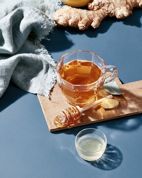 Faceted glass mug of ginger lemon tea on a wooden board with honey, featuring a cool diffuse light and moody teal background for wellness and health campaigns