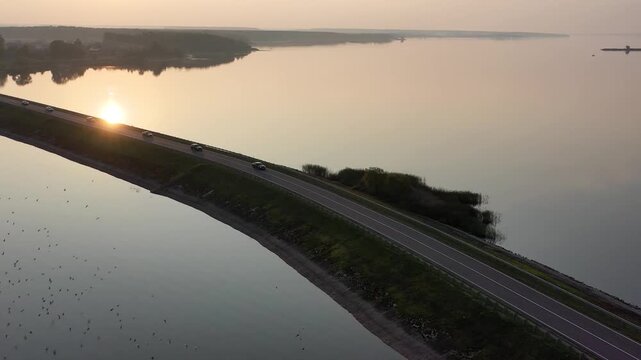 Witness the enchanting beauty of a sunrise by the river, where sunlight glimmers on calm waters and vehicles glide along the winding road. Nature comes alive in this tranquil setting.