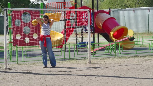 A stylish mother adjusts her hair while standing inside a soccer goal. She feels relaxed and confident on a sunny day. The colorful playground behind her brings a cheerful mood.