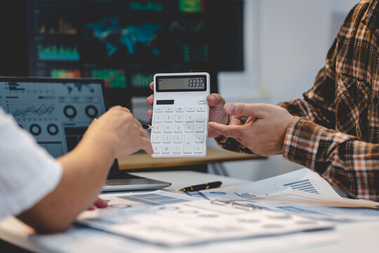 Business colleagues review charts and use a calculator at a desk, with market data screens in the background.