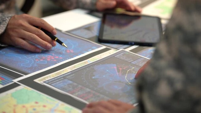 Close view of military personnel studying digital maps on tablets and paper charts during a tactical planning meeting.