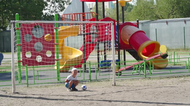A little boy squats beside a soccer ball near the goal. He smiles happily in front of a colorful playground. He feels cheerful, joyful, and ready to play outdoors.