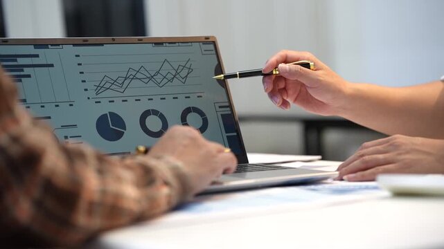 Business colleagues review financial charts on a laptop while one person points with a pen during a meeting at a desk.