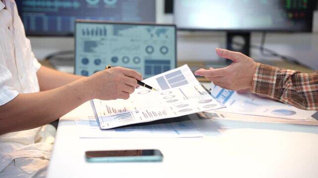 Business colleagues discuss printed charts and financial reports at a desk, with a tablet, smart phone and computer screens in a modern office.