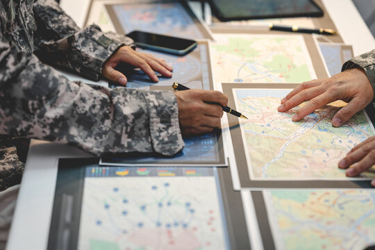 Military personnel in camouflage uniforms review printed maps and mark locations during a tactical planning meeting at a desk with digital devices.