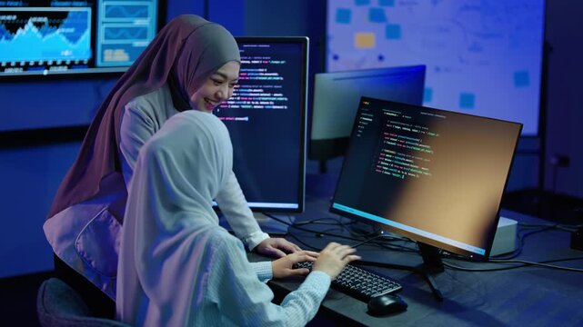 Two happy Muslim women in hijabs collaborating on programming code at a dark modern office desk. High-fiving for teamwork and coding success. Diverse women in tech and cybersecurity software concept.