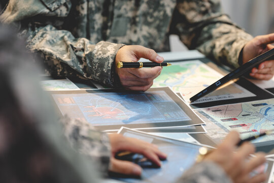 Soldiers in camouflage uniforms study maps and digital tablets during a tactical planning meeting, using a pen and touch screen for navigation and mission coordination.
